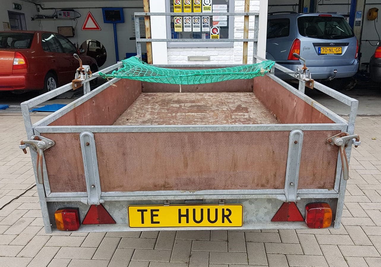 Open aanhangwagen met een gele plaat TE HUUR, op de stoep voor de geopende werkplaats.