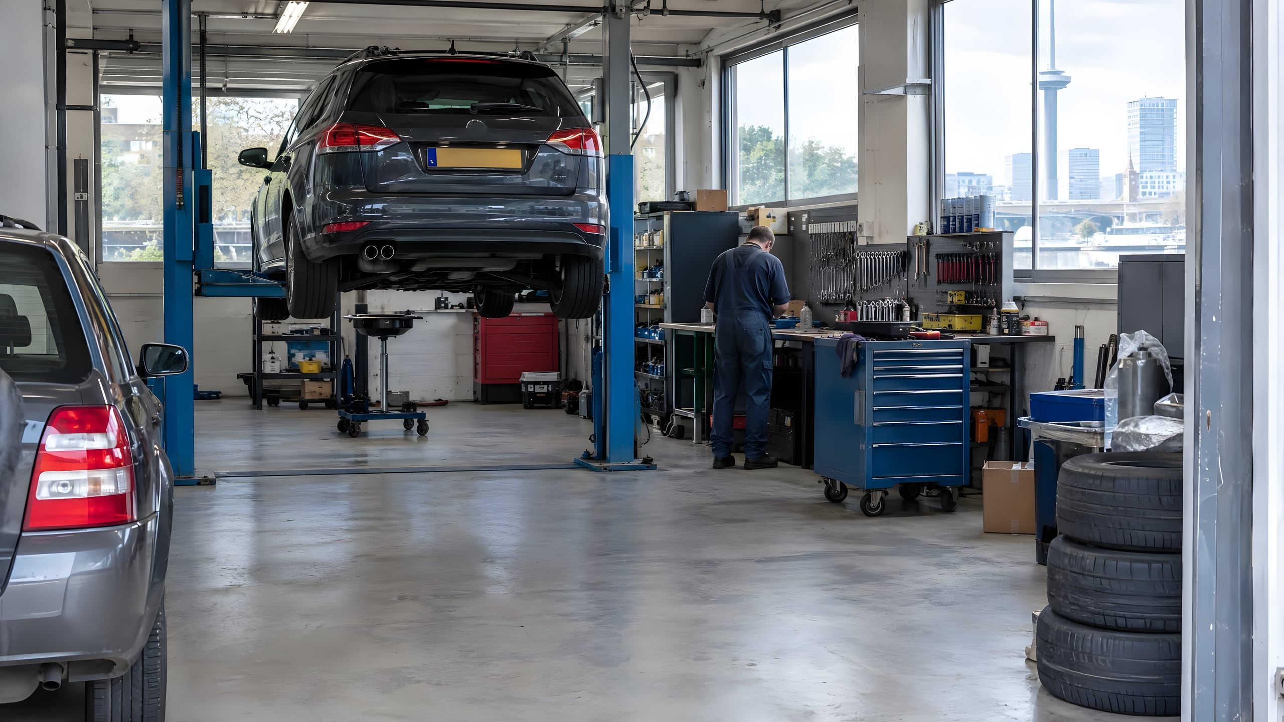Werkplaats met een auto op de hefbrug, een monteur aan de werkbank en blauwe gereedschapskasten.